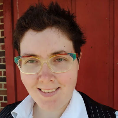 A photo of Laine Wyldling Evans, a non-binary person with spiky dark hair and light skin. They are wearing rainbow glasses, a white button up shirt, and a striped black vest.