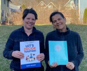 Hope Wilder, a white woman with short brown hair and Gina Price, a white woman with brown hair pulled back in a ponytail stand smiling together in front of a grassy hill with trees and houses in the background holding each of their books, Let's Decide Together and Heart to Heart.