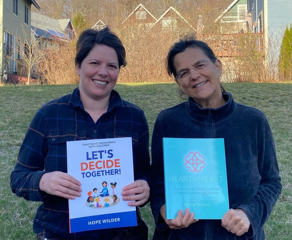 Hope Wilder, a white woman with short brown hair and Gina Price, a white woman with brown hair pulled back in a ponytail stand smiling together in front of a grassy hill with trees and houses in the background holding each of their books, Let's Decide Together and Heart to Heart.