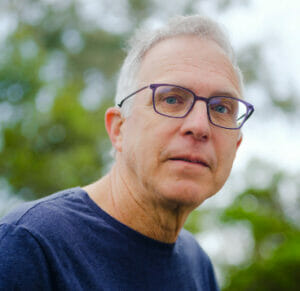Image shows David Bollier, a white man with white hair and dark rectangular glasses wearing a dark blue shirt. There are green leaves from a tree in the background.